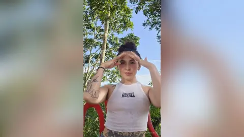 Young woman in a white ribbed tank top posing outdoors in a lush green forest setting.