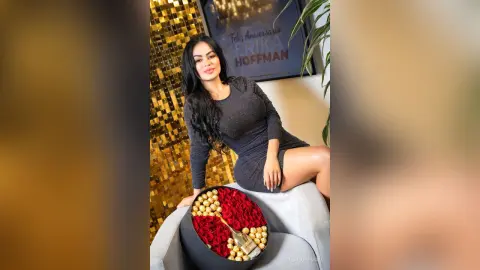 Smiling woman in a black long-sleeve dress sitting by a luxury gift box of red roses in a glamorous setting.