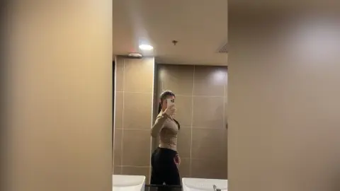 Woman in a beige long-sleeve top and black leggings taking a mirror selfie in a modern bathroom.