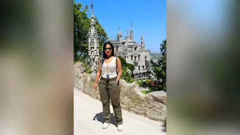 Woman in white tank top and olive cargo pants standing before a historic stone castle under a clear blue sky.
