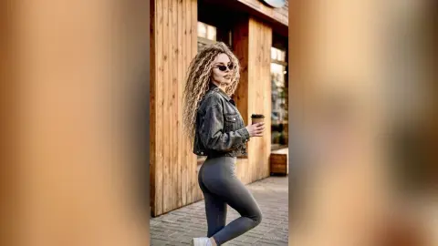 Stylish woman with long curly hair in a denim jacket and grey leggings holding coffee on a city street.