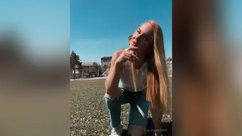 Blonde woman in a white tank top and ripped jeans relaxing on a sunny grass field under a clear blue sky.