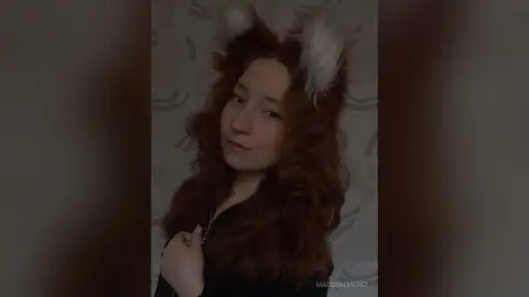 MadgeRemerez poses with her long, wavy red hair and white animal ears, wearing a black top against a patterned grey background.