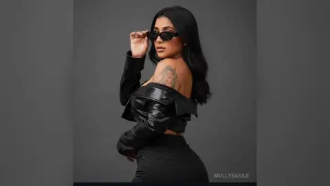 Stylish woman in black leather jacket and sunglasses posing confidently against a dark studio background.