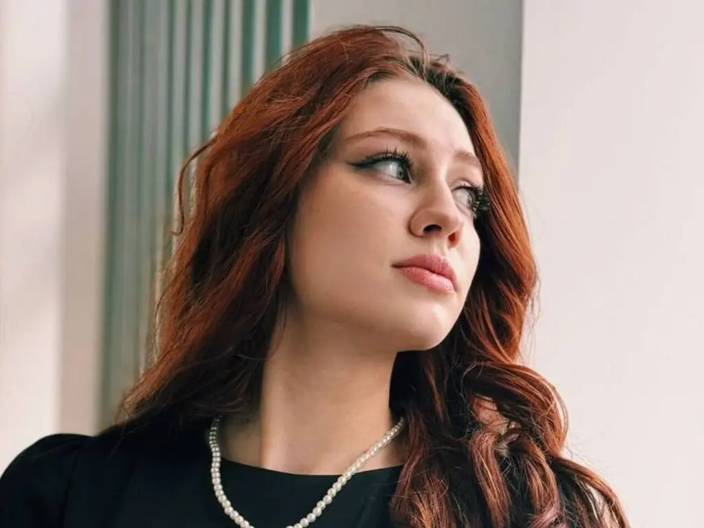 Portrait of a beautiful woman with long wavy red hair wearing a black top and pearl necklace in soft lighting.