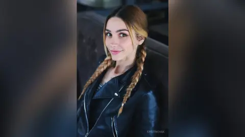 Smiling young woman with blonde braids wearing a black leather jacket in a moody indoor setting.