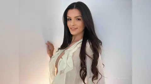 Smiling woman with long wavy hair wearing a white ruffled blouse in a bright minimalist studio setting.