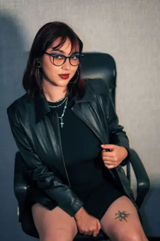 Stylish woman with glasses and dark red lipstick wearing a black leather jacket sitting in a modern office chair.