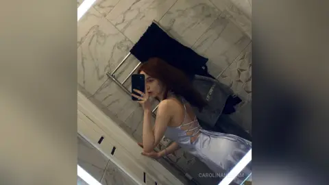 Young woman in a light blue satin slip dress taking a mirror selfie in a modern marble bathroom.