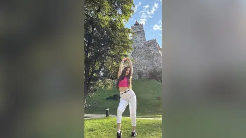 Young woman in a pink crop top and white trousers posing joyfully in front of a historic stone castle.