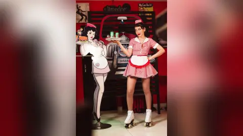 JessieLiu stands in a red and white waitress outfit and white roller skates next to a cartoon cutout of herself.