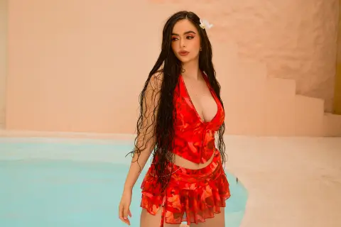 Young woman in a red floral bikini set posing by a turquoise pool in a warm, sunlit Mediterranean setting.