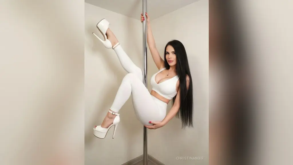 ChristinaNoir — ChristinaNoir poses on a silver pole wearing a white crop top and white leggings with white high heels, showing off her long black hair.
