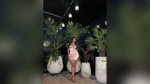 Elegant woman in a pink satin slip dress posing amidst lush green plants on a moody, dimly lit terrace.