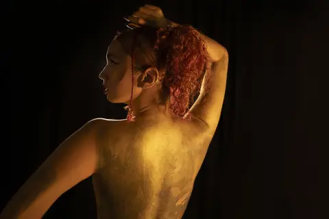 Side profile of a woman with red hair and gold body paint posing against a dark background in moody lighting.