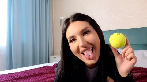 JadaJosey smiles and sticks her tongue out while holding a yellow tennis ball near her face in her bedroom.