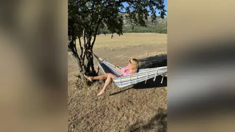 Blonde woman in a pink top relaxing in a striped hammock under a tree in a sunny meadow.