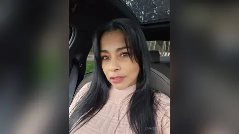 Young woman with long dark hair wearing a pink turtleneck sweater sitting in a car with a sunroof.