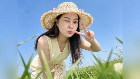CelesteQi — CelesteQi wears a straw hat and yellow sundress while posing with a peace sign in a grassy field under a blue sky.