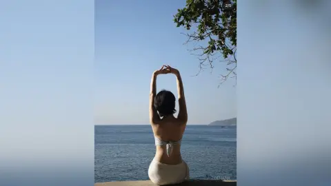 Young woman in a white bikini sitting on a pier overlooking the blue ocean under a clear sunny sky.