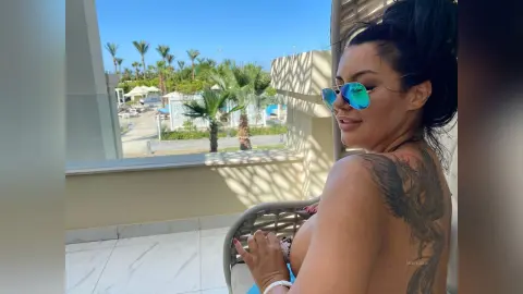 MissyJolie poses on a chair overlooking a tropical resort, showing her black hair and large bird tattoo on her back.