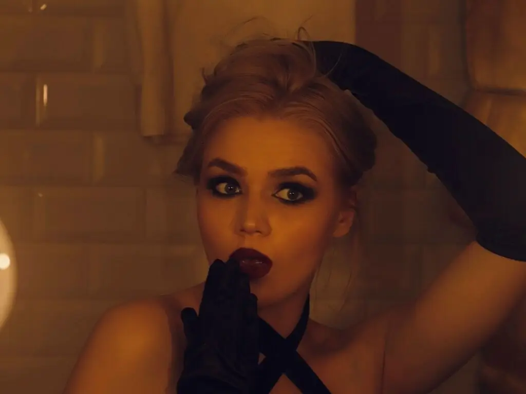 ElaineVeloz poses with her blonde hair up, wearing black gloves and dark lipstick against a tiled wall background.