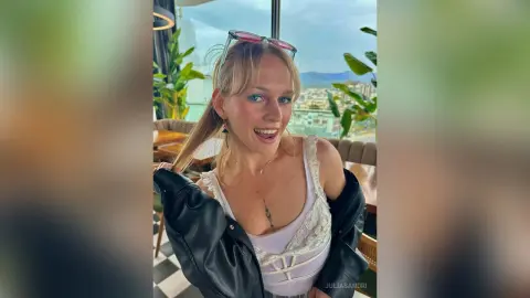Smiling blonde woman in a white lace top and black leather jacket sitting at an outdoor rooftop cafe.