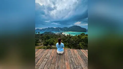 AnaMathers sits on a wooden deck overlooking a tropical coastline and mountains under a cloudy sky.