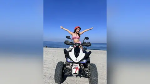 Happy woman in a pink bikini and red hat riding a white ATV on a sunny beach under a clear blue sky.