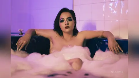 Glamorous woman with dark makeup relaxing in a bubbly bubble bath under moody purple lighting.