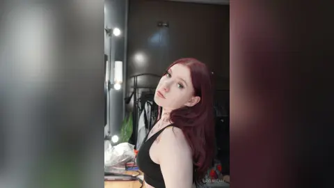 Redheaded woman in a black crop top posing in a modern bedroom setting with soft lighting.