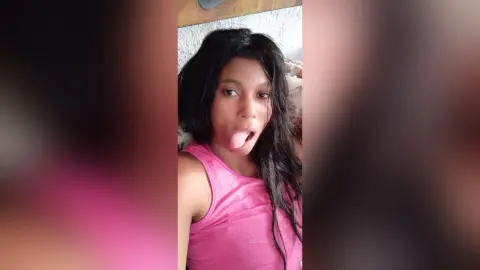 Young woman with long dark hair in a pink tank top making a playful expression in a bedroom setting.