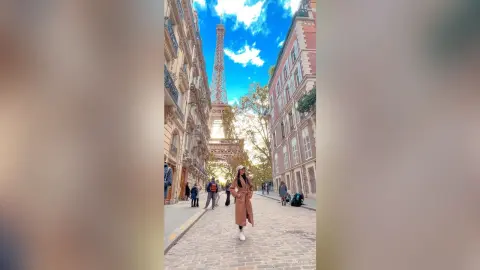 Stylish woman in a long tan coat walking on a Parisian street with the Eiffel Tower under a blue sky.