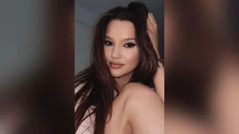 AnyaSin poses with her hand in her long brown hair, looking directly at the camera with winged eyeliner.