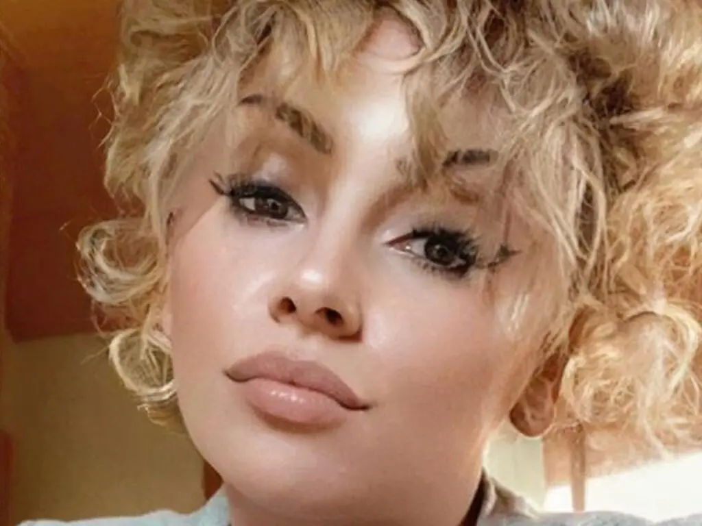 Close-up portrait of a blonde woman with curly hair and glamorous makeup looking into the camera.