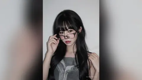 Young woman with long black hair and glasses wearing a grey sleeveless top in a minimalist indoor setting.