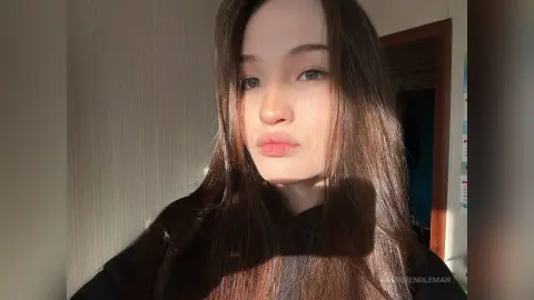 Young woman with long brown hair in a black top bathed in warm, cinematic sunlight indoors.