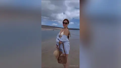 Stylish woman in a blue striped cover-up and sunglasses walking on a cloudy beach.