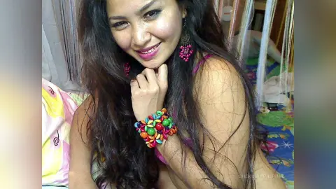 Smiling woman with long dark hair wearing colorful beaded bracelets and pink earrings in a cozy indoor setting.