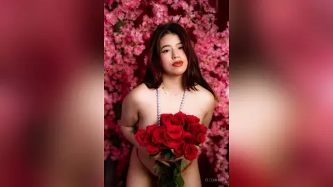 Young woman with long dark hair holding a bouquet of red roses against a romantic pink floral background.