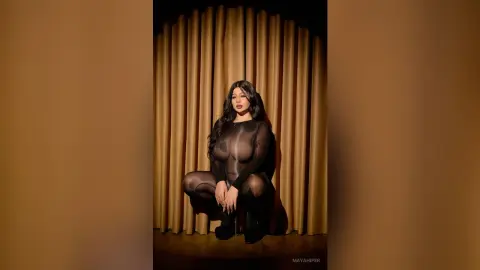 Elegant woman in a sheer black long-sleeve bodysuit posing against gold curtains in a moody studio setting.