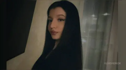 Young woman with long dark hair and winged eyeliner wearing a black top in a moody indoor setting.