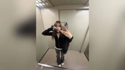 Young woman in a black zip-up hoodie and grey jeans taking a mirror selfie in a modern elevator.