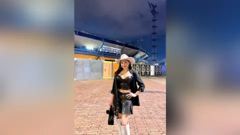 Stylish woman in a black leather outfit and white cowboy hat posing at an outdoor stadium at dusk.