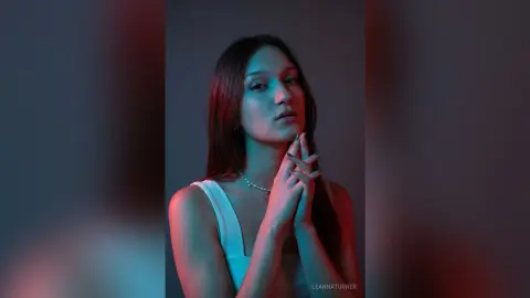 Young woman in a white tank top posing with hands clasped under moody teal and red studio lighting.
