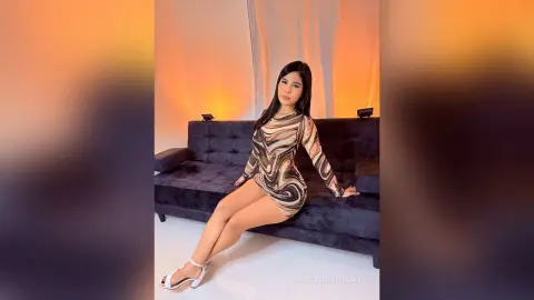 MadeleinSinclair sits on a dark couch wearing a tight, swirl-patterned long-sleeve dress and white high heels.