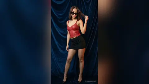 Stylish woman in a red corset top and black mini skirt posing against a blue velvet curtain backdrop.