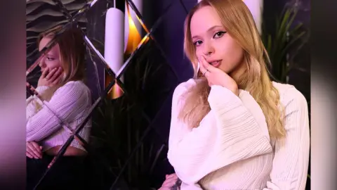LucyLoy poses with her hand on her face, wearing a white ribbed long-sleeve top in a dimly lit room.