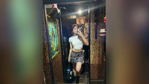 Young woman in a white crop top and plaid skirt taking a mirror selfie in a glamorous leopard print dressing room.