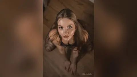 Young woman in black fishnet outfit looking up with a moody, cinematic expression in a dimly lit room.